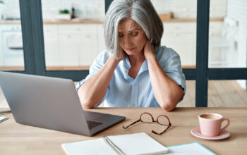 Tired stressed old mature business woman suffering from neckpain working from home office sitting at table. Overworked senior middle aged lady massaging neck feeling hurt pain from incorrect posture.